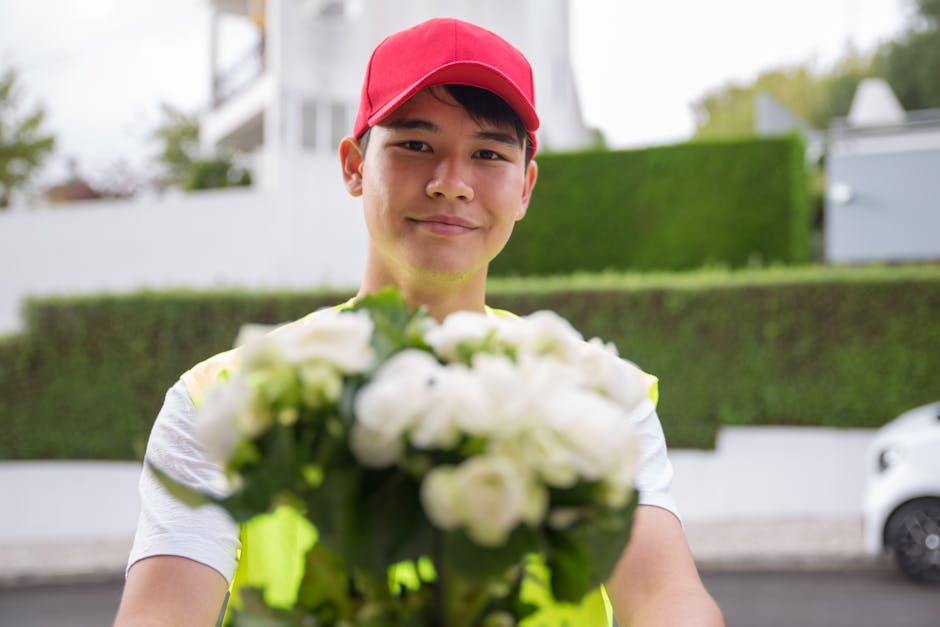 A young male florist with light skin, dark hair, and a friendly expression, wearing a bright red cap and a high-visibility vest, holding a bouquet of fresh white roses and green foliage towards the ca
