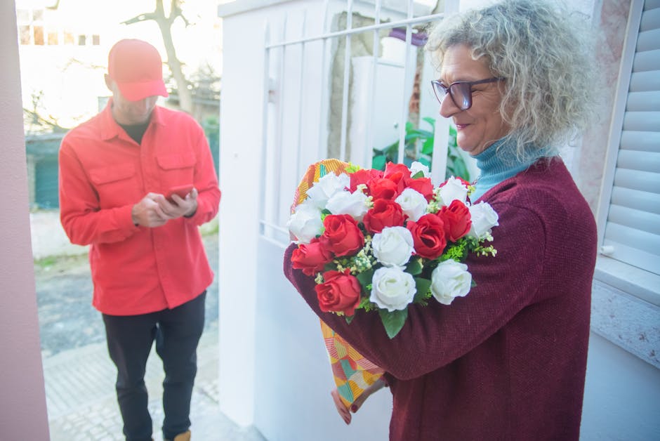 A woman with curly gray hair and glasses holds a vibrant bouquet of fresh red and white roses, wrapped in patterned paper, as she prepares to deliver or receive the floral gift. She is wearing a maroo