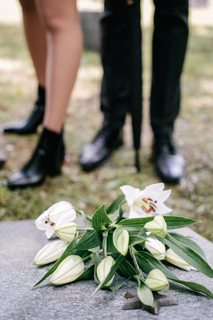 A close-up of a floral arrangement featuring white lilies with open petals revealing their reddish-brown stamens, alongside several unopened lily buds and long, slender green leaves. The flowers are p
