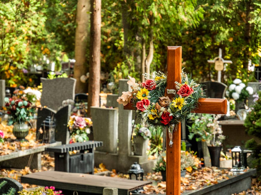 A wooden cross adorned with a floral arrangement consisting of red roses, yellow daisies, beige lilies, and green foliage, displayed at a cemetery. Surrounding the cross are various gravestones and me