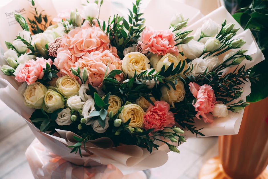 A floral bouquet arranged with soft pink carnations, creamy white roses, and pale yellow roses, accented by dark green fern leaves and eucalyptus. The flowers are carefully wrapped in cream and white 