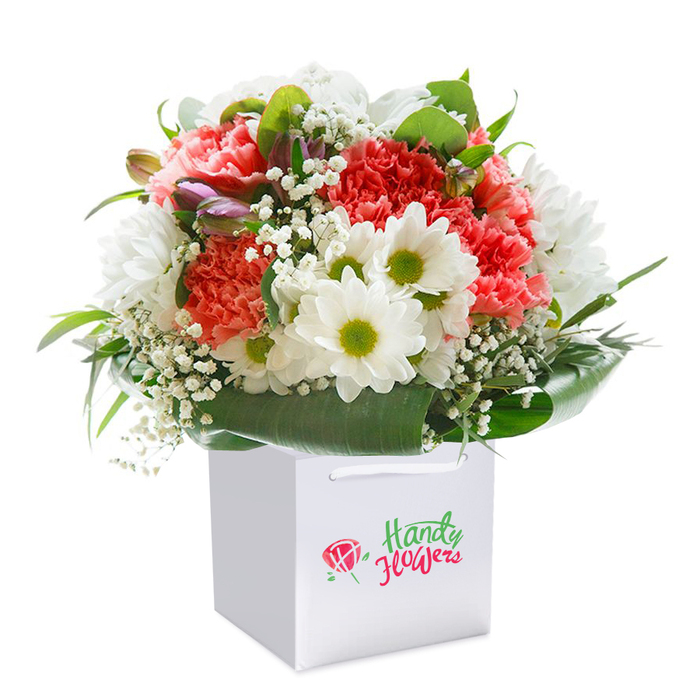 Set against a flawless white backdrop, a luxurious floral arrangement stands out in a crisp, square white gift bag-a familiar sight for Garston locals eager for fresh blooms delivered to their doorsteps. The bouquet's heart consists of lush coral and peach carnations, each petal undulating in softly frilled layers, exuding an inviting warmth and gentle fragrance reminiscent of early summer in Garston's leafy gardens. Sprinkled among these are cheerful white daisies with distinctive yellow-green centers, their pristine petals bringing a note of unclouded joy that's perfect for brightening homes near Garston station or bustling Watford Road. Sprays of white baby's breath weave a delicate lattice throughout, while slender purple-tipped buds-possibly alstroemeria-add depth and an element of surprise. The composition is encased in glossy, deep green foliage with broad leaves that curve elegantly around the base, reminiscent of the abundant hedgerows bordering Garston's many parks. The bouquet is expertly shaped into a generous, rounded form and rests neatly within the gift bag, whose durable white ribbon handles make for graceful presentation or easy collection by drivers making same-day deliveries around Garston. The carefully balanced textures and hues speak to the artistry of local florists, making this the ideal floral gift for joyful milestones-a birthday, anniversary, or simply a heartfelt expression for a friend on North Approach. This presentation captures not only freshness, but the unique blend of charm, care, and skill that defines flower delivery in Garston.