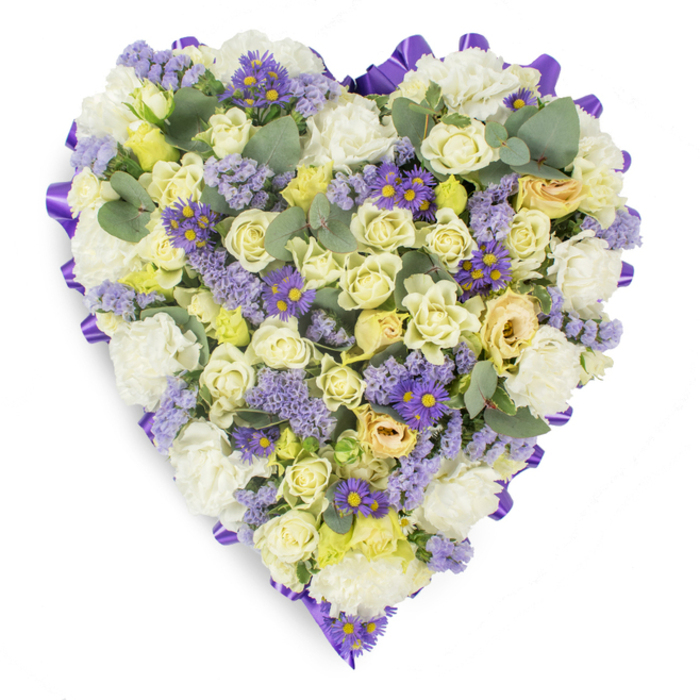 A carefully fashioned heart-shaped memorial arrangement captured against a neutral white background, showing dense, intentional floristry that reads both refined and tender. The principal components are cream and pale yellow roses, clustered tightly to form a plush, luminous centre; their soft gradients and occasional pale-green tints bring quiet visual depth and a sensation of gentle warmth. Interwoven with the roses are large, frilled white carnations that lend traditional sympathy character and soft architectural volume, creating a reassuring, cloud-like foundation. Scattered amid these lighter tones are delicate sprays of lavender statice and smaller, vibrant purple daisy-like flowers with unmistakable yellow centres-elements that add texture, richer hue, and subtle focal points for the eye to travel across the heart. Softly rounded eucalyptus leaves in blue-green hues appear throughout, offering cool contrast, a hint of silvery sheen, and a balancing, organic rhythm to the composition. The entire form is outlined by a continuous, rich purple satin ribbon folded into loops at the edge, providing a polished border and elevating the overall presentation to a formal tribute. The arrangement conveys a restrained palette of pastel and purple tones that feels meditative and respectful-suitable for funeral chapels, memorial services, or graveside placement in Garston and neighbouring communities. This piece exemplifies traditional sympathy floristry with a contemporary touch: dense, tactile blooms arranged for visual serenity and the quiet expression of enduring affection and remembrance.