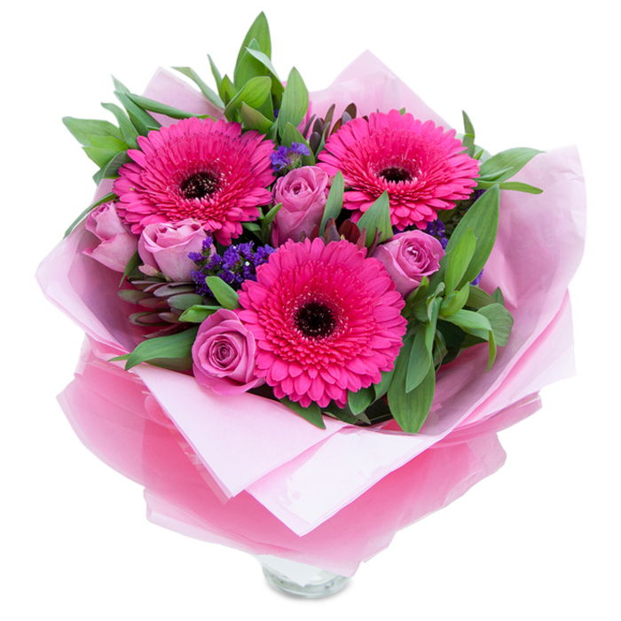 I imagine placing this bouquet on a bright windowsill in a Garston home where its colours can lift the room: three exuberant fuchsia Gerbera daisies dominate the eye, their long radiating petals surrounding striking dark centres that provide bold contrast and a playful focal point. Complementing them are tightly budded roses in a soft lavender-pink or dusty-rose, their layered petals catching light with a subtle sheen and promising to open further over the next few days. Interspersed among the main blooms, little clusters of deep purple statice add pops of texture and a slightly papery note that echoes wildflower meadows. The greenery-broad, verdant leaves that read as salal or eucalyptus-frames the arrangement with garden-fresh richness and a hint of fragrant leafiness. The floristry finish is intentional: an inner wrap of pale pink tissue paper cradles the stems, while an outer layer of translucent, crinkled fuchsia-pink cellophane lends a delicate sparkle and reinforces the bouquet's pink theme. The visible top rim of a clear glass vase at the base suggests this is a ready-to-display piece, ideal for immediate enjoyment. Scent is soft and approachable: roses deliver a light perfume, gerberas a subtle, green sweetness, and the foliage gives a clean, slightly menthol edge. Perfect for birthdays, Mother's Day, anniversaries, or a thoughtful 'just because', this bouquet feels both celebratory and intimate - the kind of arrangement a Garston florist would craft to bring a neighborly warmth after a walk past the canal or a visit to the local market.