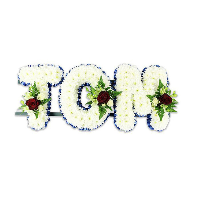 This personalised funeral tribute spells TOM in large, sculpted letters completely covered in tight clusters of small white chrysanthemum-like blooms, their petals overlapping to create a plush, tactile plane. Each letter's perimeter is crisply edged with a bright royal-blue ribbon, which provides clear visual definition and a respectful pop of colour against the white massing. Placed thoughtfully within the lower centre of each character is a composed spray: a solitary deep-red rose stands as the central emblem of love and respect, surrounded by tiny creamy spray roses and fresh green foliage including fine fern fronds that add depth and a soft, feathery texture. The contrast between the pristine whites, the saturated reds, and the fresh greens is deliberate and composed, designed to read well from a distance while also inviting close inspection. A discreet dark-green or black metal support bar is visible behind the letters, evidencing the practical care taken to display the piece upright at a memorial service. Soft, even lighting accentuates the dimensionality of the arrangement without introducing harsh shadows, and the stark white background keeps attention solely on the floral craftsmanship. As a Garston-based florist familiar with local funeral customs, I would recommend this sort of bespoke name tribute for wakes held near the town's chapels or community centres; it is a solemn, refined expression of remembrance, handcrafted to honour a life with poise and tenderness.