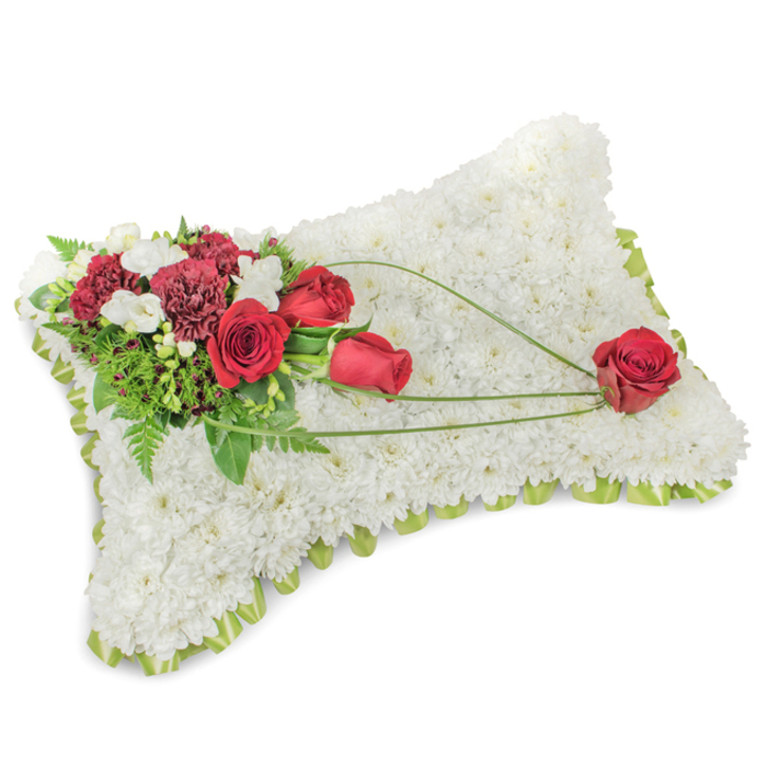 This elegant memorial cushion is composed of hundreds of compact white chrysanthemum heads, packed close to form a velvety, pillow-like surface whose pure tone conveys calm and reverence. A thin edging of light green ribbon or soft foliage peeks out along the perimeter, providing a refined outline that keeps the shape neat and intentional. Positioned on the upper left a lavish cluster of deep red roses and dark burgundy carnations commands attention; their rich, velvety textures contrast strongly yet harmoniously with the snow-white field. Interspersed among those primary blooms are small white accent flowers-likely freesias or spray chrysanthemums-that add twinkling points of light and delicate scent. Layers of green leaves and feathery fern fronds cradle the spray, contributing depth and a fresh, natural undertone. From that corner the eye follows a trio of slender green blades or bear-grass, arcing gracefully across the cushion toward the lower right where a single, perfect red rose rests as if gently placed there by design. The composition reads as both crafted and tender, lit in soft daylight that brings out petal sheen and subtle shadows, and the overall fragrance is clean and reassuring. Locally minded and compassionate in tone, this floral pillow is the kind often chosen by families around Garston and neighbouring streets to express condolence at funeral services, wakes, or memorial gatherings, combining traditional symbolism with careful florist craftsmanship to honour a life with dignity and quiet love.