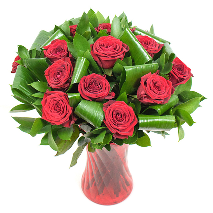 This composition showcases roughly a dozen rich, crimson roses, each blossom opened to reveal densely layered, velvety petals that curl inward in precise spirals. The colour is saturated and deep, a passionate red that darkens gracefully toward the petal bases while the tips catch a soft, flattering light. Interspersed between the rose heads is a lavishing of deep green foliage: smaller, glossy leaves create a plush backdrop while larger, sculpted leaves have been folded and curled into cylindrical arcs and loops that cradle the roses and introduce an architectural rhythm to the round bouquet. The textural play between the smooth, shiny greens and the matte, velvety petals makes the arrangement feel both tactile and composed. The stems are contained within a tall, slender ruby-red glass vase whose translucent body is softly ribbed or twisted, throwing slender highlights up its neck and echoing the verticality of the flowers. The vase's rich red hue harmonises with the roses while the pure white background strips away distractions, letting the arrangement read as a focused statement-ideal for anniversaries, Valentine's gestures, or an elegant table centre. I imagine the scent as a clean, classic rose perfume that fills a small room, the vase warm in the hands as it's carried across Garston's neighbourhoods toward Garston Dock or nearby Speke. This bouquet's combination of timeless roses and modern leaf-folding is a favourite for clients seeking a sophisticated, heartfelt floral message.