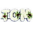 The photograph captures a traditional name tribute designed for a funeral or memorial, with the word TOM spelled out in large, three-dimensional capital letters made entirely from white chrysanthemums. Each letter is densely packed with these small, daisy-like blooms, their petals forming a smooth, plush surface that looks soft to the touch and perfectly even from edge to edge. Framing the outside of the T, O and M is a bold royal blue satin ribbon, carefully folded into neat pleats that trace every straight line and rounded corner, giving the tribute a refined outline and a sense of formal dignity. At carefully chosen points on each letter - high on the left of the T, along the rounded side of the O, and towards the upper right of the M - compact sprays of accent flowers break through the white. These feature deep crimson red roses, some in tight buds and others open and full, accompanied by smaller white or cream spray roses and slender white, freesia-like blooms. Their rich colour is enhanced by a surround of mixed greenery, including soft fern fronds and polished leaves that add texture and depth, much like the layered greenery seen in mature Garston gardens. The three letters sit side by side on a slim, dark frame, which supports them discreetly and suggests the tribute is ready to rest alongside a coffin, near a lectern, or at a graveside. Overall, the arrangement feels both strong and tender, a bespoke sympathy flower design that spells Tom's name clearly and beautifully, offering a serene and respectful focal point for those gathering to say goodbye in and around Garston.