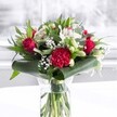 A striking red and white bouquet stands proudly in a clear cylindrical glass vase, its fresh green stems visible beneath the water like a neat forest of colour, as if just arranged in a Garston florist's studio. Deep red roses, their velvety petals beginning to unfurl, form the romantic heart of the design, while a richly ruffled crimson carnation sits low at the front, creating a textured focal point that immediately draws the eye. Around them, white alstroemeria with slender, speckled petals and creamy chrysanthemum-style blooms introduce softness and calm, their pale tones glowing in the gentle daylight that filters across the image. Tucked between these larger flowers are sprays of tiny white gypsophila, sprinkling the bouquet with a cloud-like lightness, almost like a dusting of fresh snow over red velvet. Curled, glossy dark green leaves are wrapped cleverly around the base of the arrangement, forming an elegant collar that both frames and supports the flowers, while assorted foliage with eucalyptus-like tones and pointed leaves adds depth, movement, and a layered tapestry of green. The arrangement rests on a pristine white surface, in front of a softly blurred pale background that could be a bright Garston window or a light wall, giving the impression of a calm living room or office along St Mary's Road or near Garston Park. The lighting is even and flattering, catching the highlights on each petal and leaf, creating a sense of freshness and celebration. This bouquet feels perfectly suited to anniversaries, birthdays, or a thoughtful delivery to a Garston home or workplace, sharing joy, admiration, and warm affection through classic colour and careful craftsmanship.