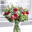 A generously full bouquet of deep red roses and matching red carnations stands proudly in a clear glass vase on a bright white tabletop, the whole arrangement glowing in soft daylight filtering through a large window. Each rose in this Garston-inspired design shows off velvety petals, some fully open in rich crimson spirals, others still in tight buds that promise more colour in the days ahead. Nestled between the roses, ruffled carnations echo the same passionate tone of red, adding texture and volume so the bouquet feels plush and abundant, like something you might discover in a favourite café near Garston Park. Threaded through the bold reds are sprays of crisp white alstroemeria, a mix of closed buds and half-open trumpets, their petals tipped with the faintest suggestion of green that catches the light. Tiny clusters of baby's breath, or gypsophila, form a soft, cloudlike veil around the brighter blooms, giving the design a romantic haze. The flowers are cradled by large, glossy green leaves that create a structured collar at the base, while finer stems of lighter greenery weave gracefully upward, adding movement and depth. Through the sides of the simple cylindrical vase, you can see neatly trimmed stems standing in clear water, reinforcing the feeling of just-delivered freshness you'd expect from a local Garston florist. Behind the bouquet, the outdoor view is blurred and almost monochrome, so the reds, whites, and greens appear even more vivid and celebratory. The mood is joyful and heartfelt, perfect for a romantic anniversary, a Mother's Day surprise, or simply sending warm wishes to someone special in and around Garston.