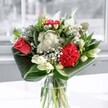 A contemporary floral arrangement in a clear glass vase is captured in soft daylight, standing on a simple surface near a bright window, as if placed in a calm Garston living room just off St Mary's Road. At the heart of the bouquet, a single coral-red rose and a rich crimson carnation create an immediate focal point, their velvety petals layered and plush, suggesting both passion and warmth. Around them, clusters of pure white chrysanthemums and small daisy-like blossoms drift gently, their fine petals giving a cloud-like softness that contrasts with the bold red tones. Creamy white lilies sit mostly in bud, their long green stems and gently tapered forms adding height and structure, hinting at blooms still to come and the quiet promise of new beginnings. Deep emerald foliage is folded and curved around the base, its glossy leaves forming sweeping arcs that cradle the flowers and give the design a modern, polished edge often seen in boutique arrangements across Garston and nearby Aigburth. The clear glass vase reveals clean stems and fresh water, catching reflections of the light that streams in from the window. The overall impression is of freshness and serenity, as though a cool morning breeze from nearby parks has been gathered into flowers. This bouquet feels ideal for celebrating a special milestone, offering congratulations, or simply bringing a gentle lift to an ordinary afternoon at home in Garston, with the subtle scent of lilies and greenery imagined as drifting softly through the room.