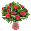 A rich, rounded bouquet of a dozen or more deep crimson roses fills the frame, each bloom opened to a velvety, spiralled perfection that reads as classic romance. The petals are a wine-dark red, their satiny texture catching soft, even light so the subtle ridges and inner folds are clearly visible. Nestled between the rose heads is an abundant cloak of dark green foliage: flat, glossy leaves form a dense backdrop while larger, emerald-green leaves are expertly folded, rolled and curled into sculptural loops that cradle individual blooms like green ribbons. Those architectural leaf forms-reminiscent of Aspidistra and polished Ruscus-add modern structure and a tactile contrast to the softness of the roses. The whole composition sits in a tall, translucent ruby-red glass vase with a faint ribbed twist that echoes the vertical lines of the stems and reflects highlights along its curved silhouette. Placed against a clean white background, the intense reds and lush greens pop with clarity, isolating the arrangement as an elegant centerpiece or a thoughtful, passionate gift. Imagine the gentle, classic perfume of fresh roses rising from the cluster and the reassuring weight of the glass vase when carried from a Garston doorstep; this is a design I would recommend for anniversaries, Valentine's Day, or any celebration that calls for timeless romance. As a local florist familiar with Garston village stalls and the Mersey's softer evening light, I pictured this arrangement to read beautifully in photographs, on a mantelpiece, or as a memorable delivery for a loved one in the neighbourhood.