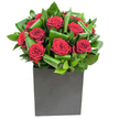 An encounter of refined modern romance: a compact cluster of deep, velvety red roses sits at the heart of this arrangement, each bloom unfurling in generous spirals that catch the eye with a velvet sheen. Broad, glossy green leaves have been carefully curled and overlapped around the roses, forming rhythmic arcs that add movement and sculptural depth without stealing the show. The composition rests inside a crisp matte black cube - a low, square vessel whose stark geometry grounds the design and amplifies the intensity of the crimson above. Lighting is bright and clear, the kind of steady studio light that brings out subtle highlights on each petal and the reflective surfaces of the foliage, so the arrangement reads as both sumptuous and contemporary. If you could reach in you would feel the silken softness of the petals against the firmer, almost waxy texture of the leaves; if you drew close, the scent would be a clean, classic rose perfume moderated by green, leafy top notes that give the bouquet a refreshing lift. This piece carries a quietly dramatic mood, equally at home as an intimate anniversary gift, a soulful apology, or a tasteful centrepiece on a modern dining table. Made with local sensibility, its understated glamour speaks well to Garston homes and offices seeking a polished, heartfelt statement. The craftsmanship is deliberate-folds, overlaps and negative space arranged to draw attention to each bloom-so the bouquet feels like a small piece of floral sculpture that invites both sight and touch, and lingers in memory long after the vase is set down.